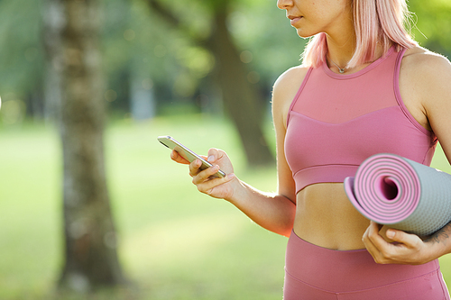 Close-up of young sporty woman in pink sports clothing holding exercise mat and looking at her mobile phone in her hand while standing outdoors