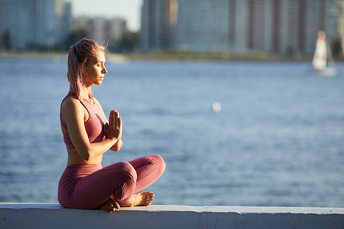 Young healthy woman sitting with eyes closed in lotus position and meditating in the city with sea in the background