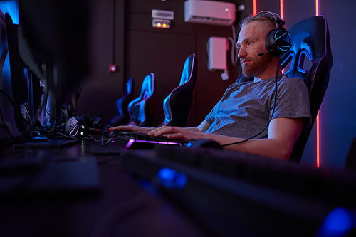 Young gamer in headphones typing on computer keyboard and looking at computer monitor while sitting on computer chair