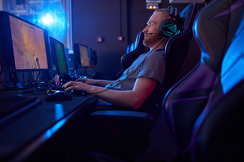 Young bearded man in headphones sitting in gaming chair and playing in computer game on computer in dark room