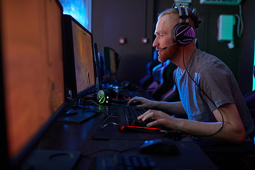 Young serious man wearing gaming headphones sitting in front of computer monitor and concentrating on his game