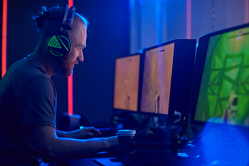 Young man in gaming headphones looking seriously he sitting in front of computer monitors and playing game in dark computer class