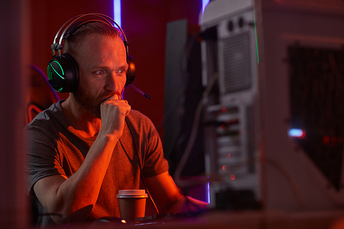 Young serious computer programmer in headphones looking at computer monitor while sitting at his workplace at office