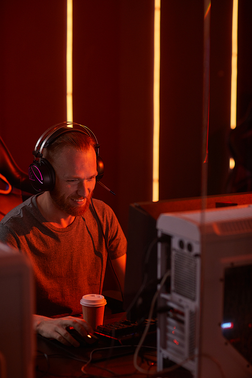 Young bearded man in headphones sitting at the table in front of computer and smiling during his work