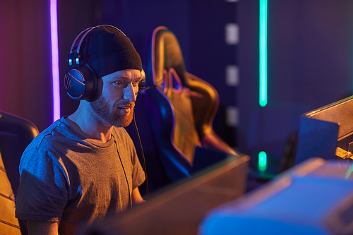 Young bearded man in hat and in headphones sitting in front of computer monitor and developing new program