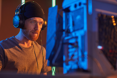 Young bearded man in hat and in headphones concentrating on his work he sitting at his workplace and looking at monitor