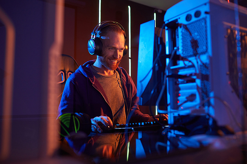 Young bearded man in headphones working on computer and smiling while sitting at his workplace in dark office