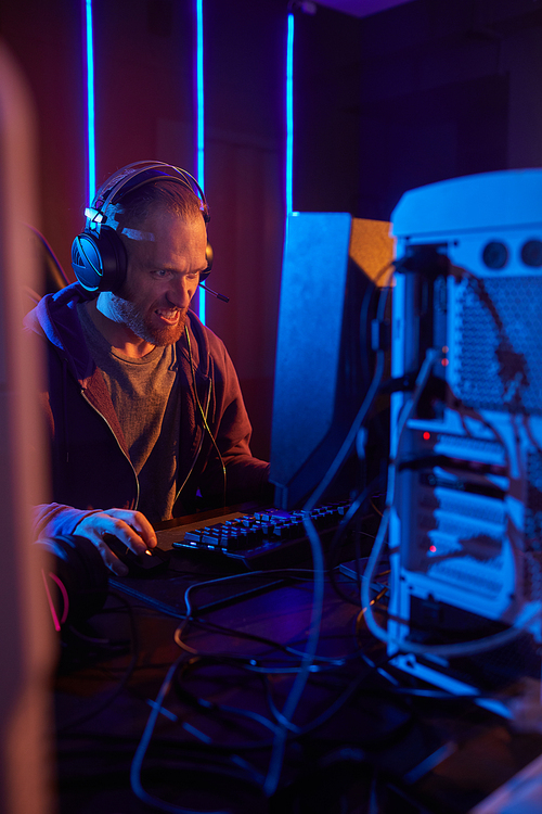 Young computer programmer in headphones looking very nervous he sitting at the table and working on computer in dark office