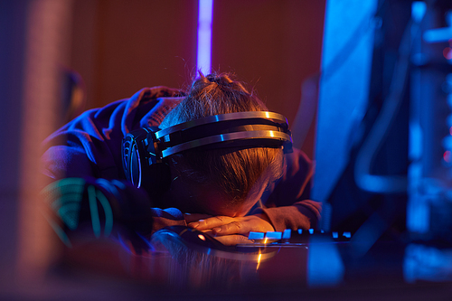 Young computer programmer in headphones sleeping at his workplace in front of computer in dark office