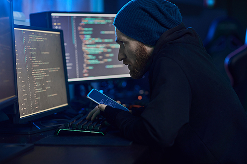 Computer hacker in dark clothing sitting in front of computer monitors and using his mobile phone to break into the system