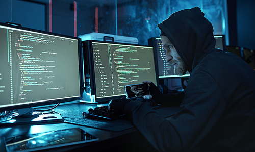 Young man in hoody using mobile phone and reading the information from computers in dark office