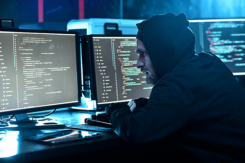 Young computer hacker in hoody looking at computer monitor and breaking the software in dark office