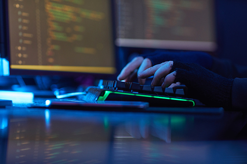 Close-up of man wearing black gloves typing on computer keyboard at the table in dark office