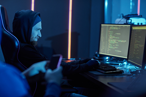 Young man in black clothing typing on computer keyboard and smiling he stealing the information from computers together with his partner