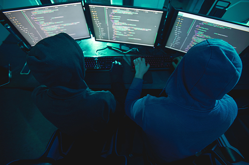 Rear view of two computer hackers in hoodies typing on keyboard and breaking the computer system while sitting in dark office