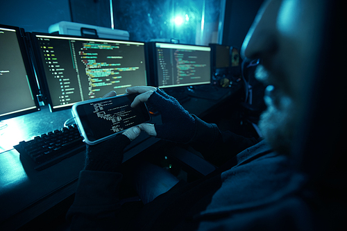 Close-up of computer hacker looking at computer monitor and guessing the password using his mobile phone in dark room