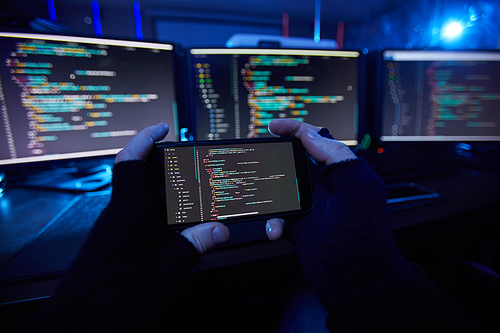 Close-up of computer hacker in black gloves holding mobile phone and taking a photo of computer software on it in dark office