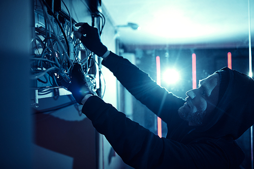 Young man in hoody standing near the box on the wall and working with cables he repairing the electricity system