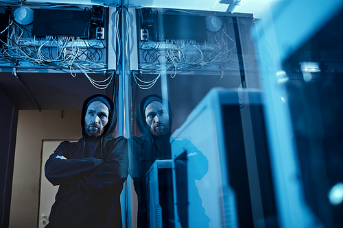 Serious computer programmer in black clothing standing with arms crossed with computer system with cables over him in dark office