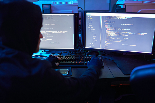 Rear view of computer hacker wearing hoody sitting at the table in front of computer monitors and breaking the computer codes in dark office