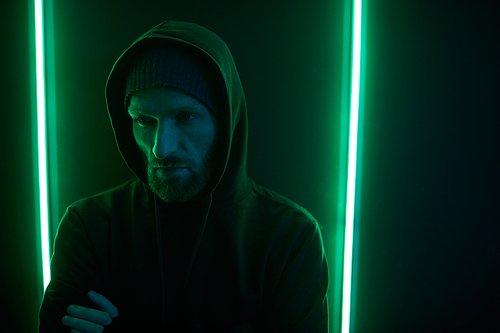 Young bearded man wearing black hoody shirt standing with arms crossed against the wall with green light