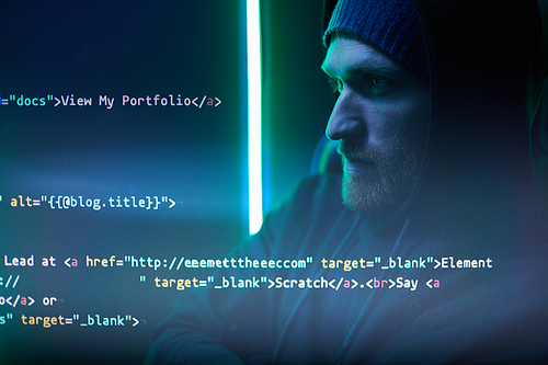 Computer hacker in hoody sitting in front of computer monitor and reading symbols on it in dark room