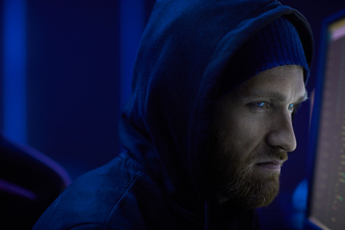 Serious computer hacker in black hoody shirt looking at computer monitor with serious expression of the face in dark room