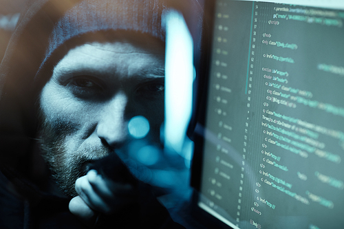 Portrait of young bearded computer programmer looking at camera near the computer monitor with software in dark office