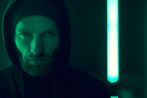 Portrait of serious bearded gangster wearing black hoody shirt looking at camera with green lights in the background