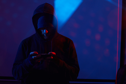 Image of computer hacker in black clothing standing and using his mobile phone in dark room