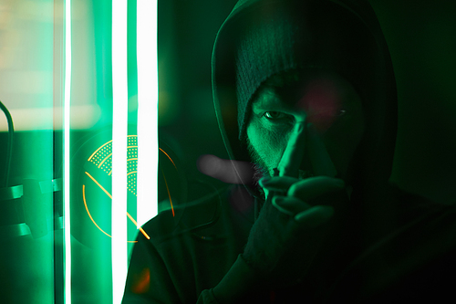 Portrait of computer hacker in black hoody looking at camera in dark room with green light in the background