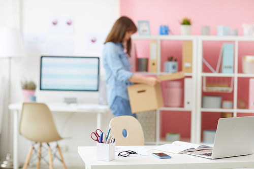 Focus on office desk with modern devices and planner, young woman carrying cartoon box in background