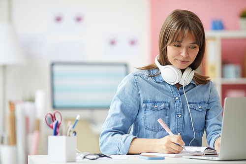 Content hipster woman in denim shirt sitting at table and making notes in organizer while working with analytical information
