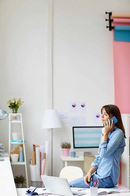 Pensive photo studio administrator in denim outfit sitting on table and calling client on cellphone