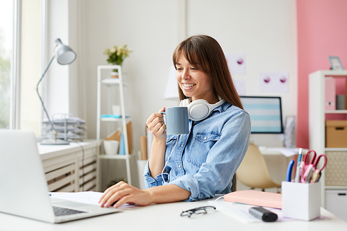 Cheerful attractive young woman with white headphones on neck sitting at table and drinking coffee while viewing online resources