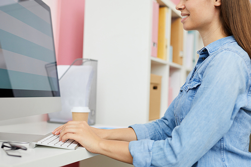 Close-up of content young woman in denim shirt sitting at table and typing on computer keyboard while creating website design