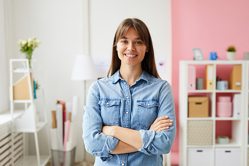 Portrait of smiling confident young lady with brown hair wearing denim shirt standing in office with pink interior