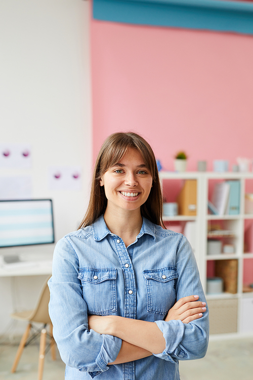 Positive pretty young lady crossing arms on chest and standing in cute office with shelves in background