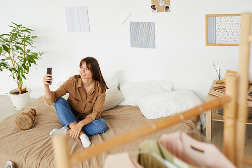 Smiling student girl with brown hair sitting on bed and having online communication via mobile app
