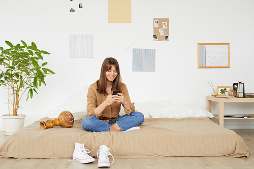 Smiling attractive young woman with brown hair sitting with crossed legs on floor bed and using smartphone in cozy bedroom