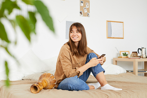 Positive young woman with bangs sitting on bed with djembe and looking away while thinking of sms