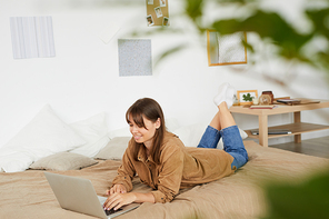 Content freelance girl with brown hair lying on bed and typing on laptop while working at home