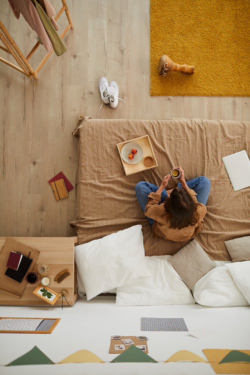 High angle view of brunette lady sitting with crossed legs on bed and drinking lemon tea while having snack in bedroom