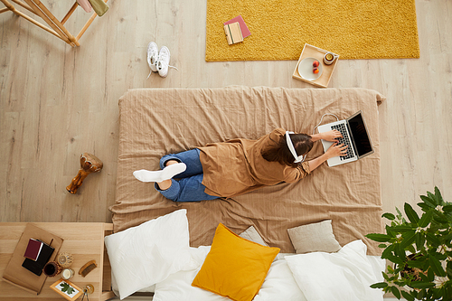 High angle view of young woman in headphones lying in bed and typing on laptop while working on research