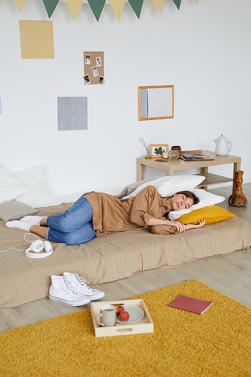 Tired young woman in casual clothing sleeping on floor bed in cozy students bedroom with scattered things