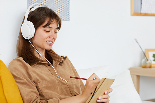 Smiling pretty girl in white headphones inspired with music making notes in sketchpad while sitting at home