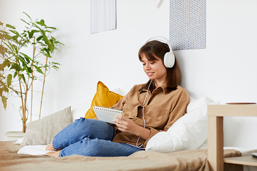 Content creative young girl in white headphones sitting on bed with comfortable pillows and making notes in organizer while planning day