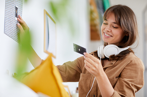 Smiling attractive young woman with brown hair looking at instant photo while decorating room with photographs