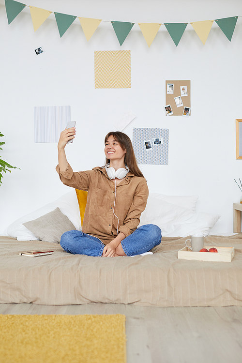 Happy excited modern girl with headphones on neck sitting with crossed legs on bed and chatting via video link