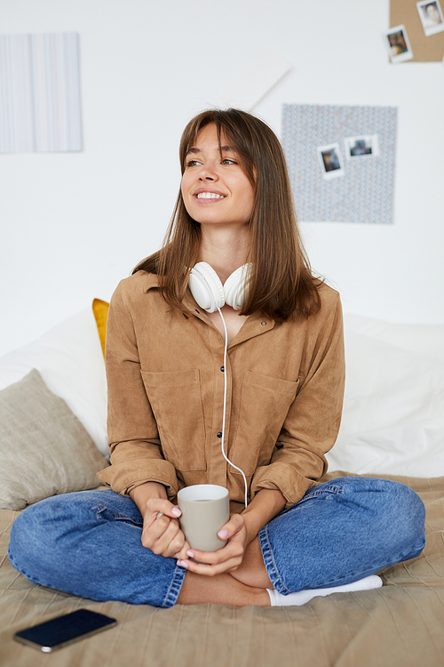 Positive calm young woman with brown hair sitting with crossed legs on bed and holding mug while resting at home